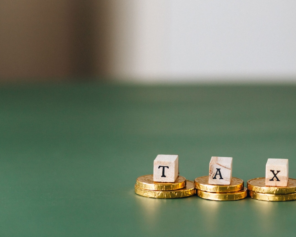 Stacked gold coins with wooden blocks spelling 'TAX' on a green backdrop.